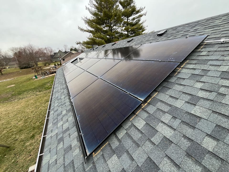 Black solar array on a gray shingle roof, with an open lawn and trees behind