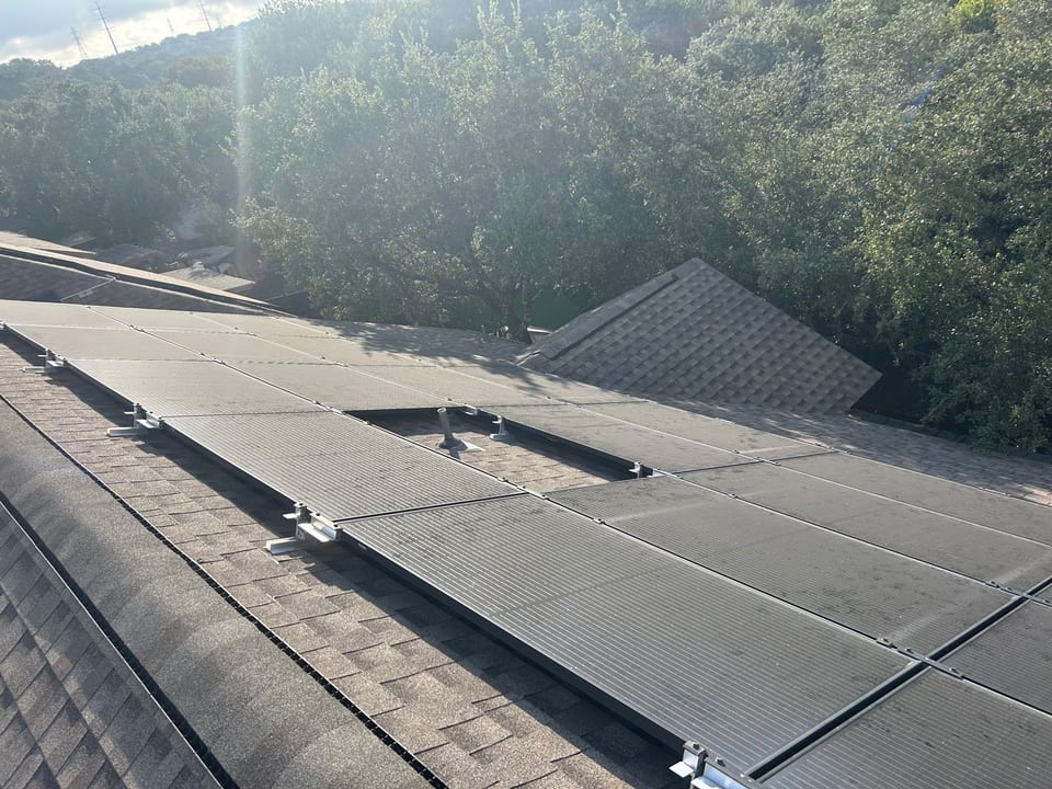 Solar array spanning a shingle roof, backlit by morning sun through the trees